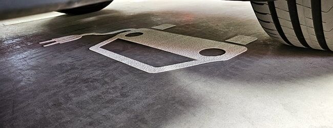 Parkplatzmarkierung in Form eines stilisierten Ladestecker-Symbols für Elektrofahrzeuge neben einem Autoreifen.