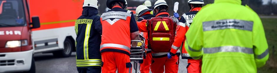 Rettungskräfte in verschiedenfarbigen Warnwesten (orange, gelb, rot) transportieren eine Trage bei einem Einsatz.
