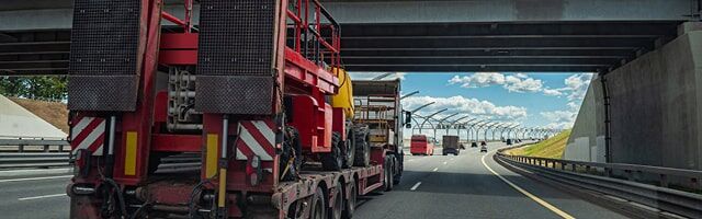 Rot-weiße Warntafeln Ein Lkw mit rot-weißen Warntafel, die Überlänge markieren, fährt unter einer Brücke hindurch, während die Sonne im Hintergrund scheint.