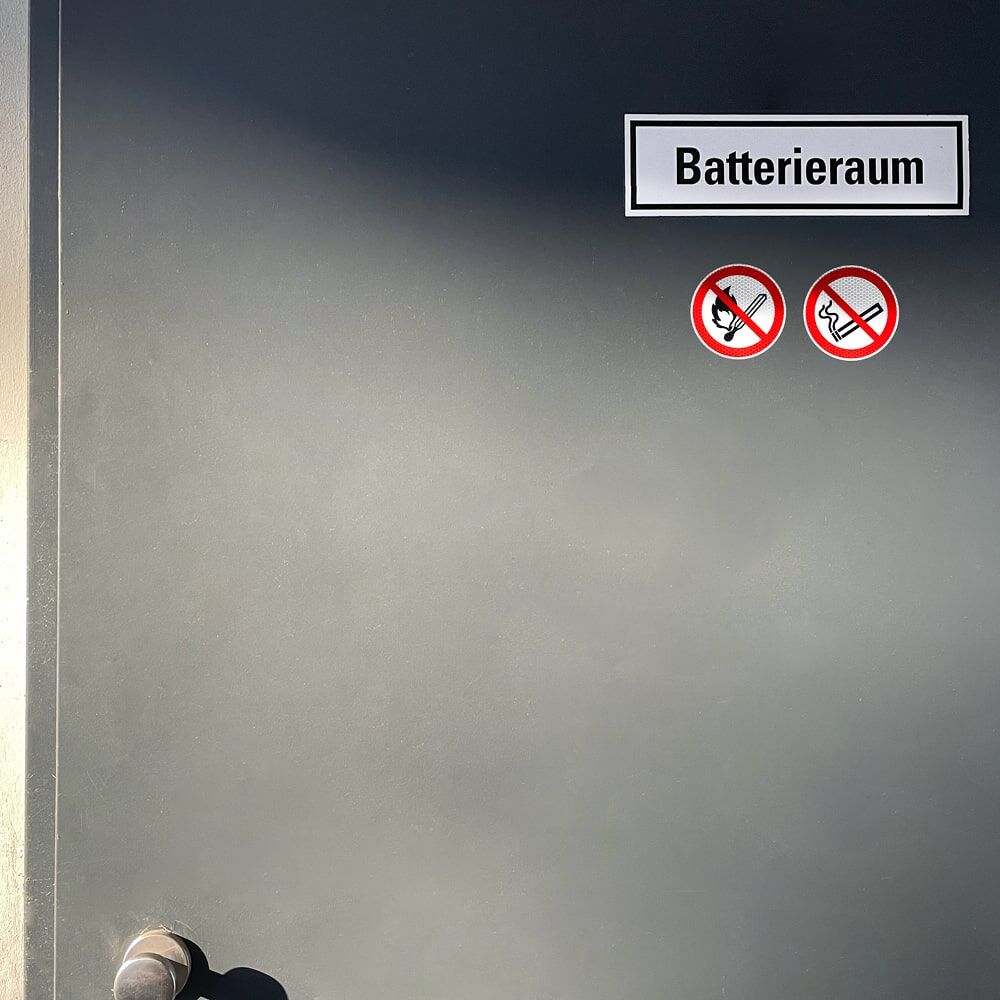 Anwendung der Aufkleber „Keine offene Flamme“ und „Rauchen verboten“ an Tür zum Batterieraum